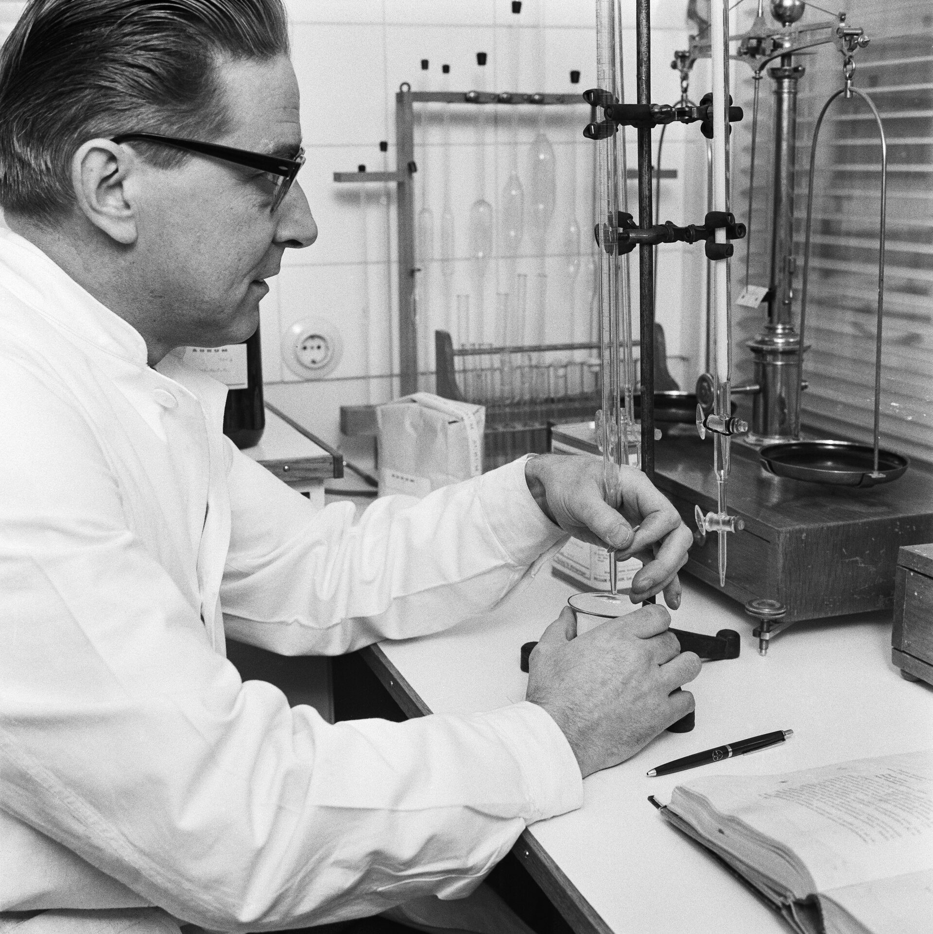 Farmacéutico con bata blanca valorando una muestra con una bureta, junto a una balanza de precisión, en 1965