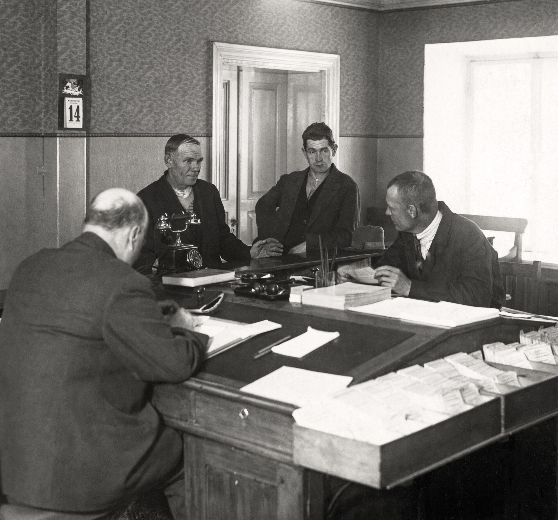 Oficina de empleo en 1931: un empleado anota en un libro frente a tres hombres que esperan, con un fichero de tarjetas sobre la mesa